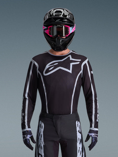 Maillot Alpinestars Fluid Apex 2026, maillot de motocross, noir et gris, avec un grand logo Astars blanc sur la poitrine et des détails graphiques gris sur les manches, porté par un pilote avec des lunettes roses et un casque noir, équipement de course tout-terrain coordonné