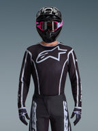 Ensemble d'équipement Alpinestars Fluid Apex 2026, maillot et pantalon de motocross noir et blanc, avec un grand logo sur la poitrine et des lignes géométriques, porté avec un casque noir et des lunettes roses, équipement de performance pour le motocross tout-terrain