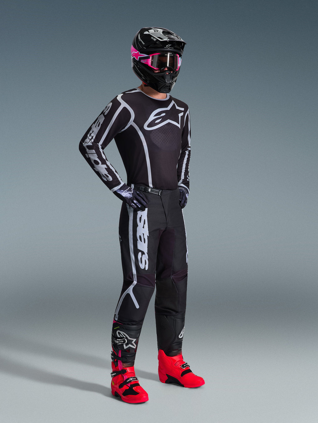 Ensemble d'équipement Alpinestars 2026 Fluid Apex, pilote de motocross portant un maillot et un pantalon noirs avec des motifs graphiques blancs et gris, casque noir avec lunettes roses et bottes de course rose vif, tenue de motocross professionnelle coordonnée