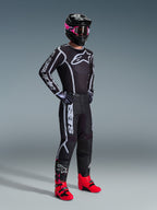 Ensemble d'équipement Alpinestars 2026 Fluid Apex, pilote de motocross portant un maillot et un pantalon noirs avec des motifs graphiques blancs et gris, casque noir avec lunettes roses et bottes de course rose vif, tenue de motocross professionnelle coordonnée