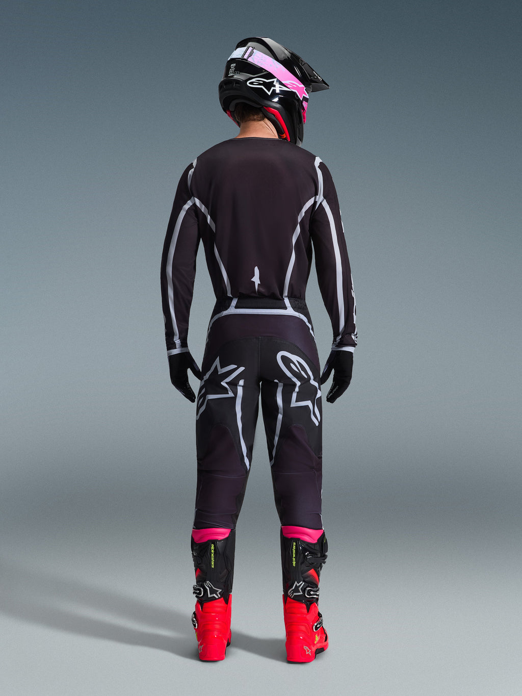Motocross rider from the back wearing the Alpinestars 2026 Fluid Apex Jersey and matching pants in Black/Gray, featuring white geometric design lines and star logos on the seat, accessorized with a black and pink helmet and red and black racing boots.