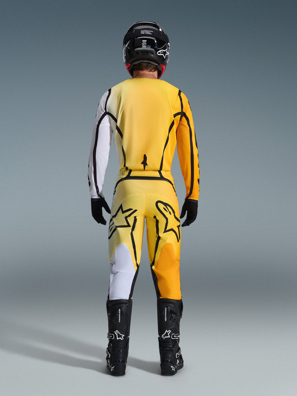 Motocrossrijder van achteren gezien in de Alpinestars 2026 Fluid Apex Gear Set, geel jersey met een witte mouw en zwarte accenten, bijpassende gele broek met grote zwarte Alpinestars-logo's op de achterzijde, zwarte helm, zwarte handschoenen en zwarte laarzen, gecoördineerde outfit voor off-road racen
