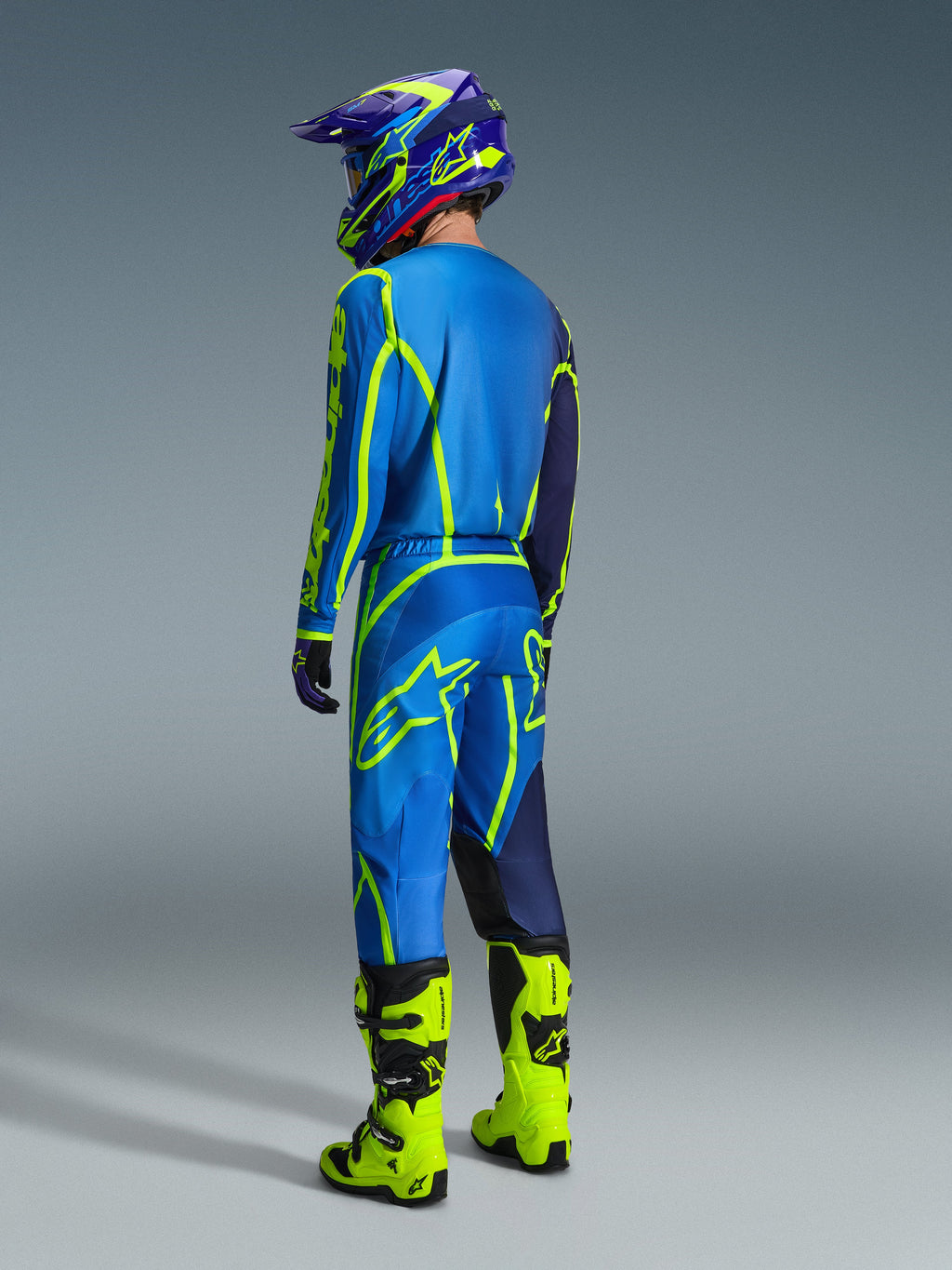 Alpinestars 2026 Fluid Apex Gear Set, Motocross-Fahrer in der Rückansicht mit blau-neongelber Ausrüstung, blaues Jersey und Hose mit neongelben Logos, neongelbe Stiefel, lila-blauer Helm, koordinierte Offroad-Rennbekleidung.