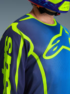 Nahaufnahme des Alpinestars 2026 Fluid Apex Trikots in Blau und Gelb Fluo, mit einem großen neongelben Astars-Logo auf der Brust, passendem Motocross-Helm und feuchtigkeitsableitender Polygewebe-Konstruktion.
