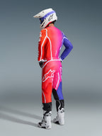 Pilote de motocross portant l'ensemble d'équipement Alpinestars 2026 Fluid Apex, présentant un dégradé éclatant de l'orange au rose et au violet avec des logos en étoile et un marquage blanc audacieux, accompagné d'un casque blanc, de lunettes bleues et de bottes blanches, vue de trois-quarts arrière en studio.