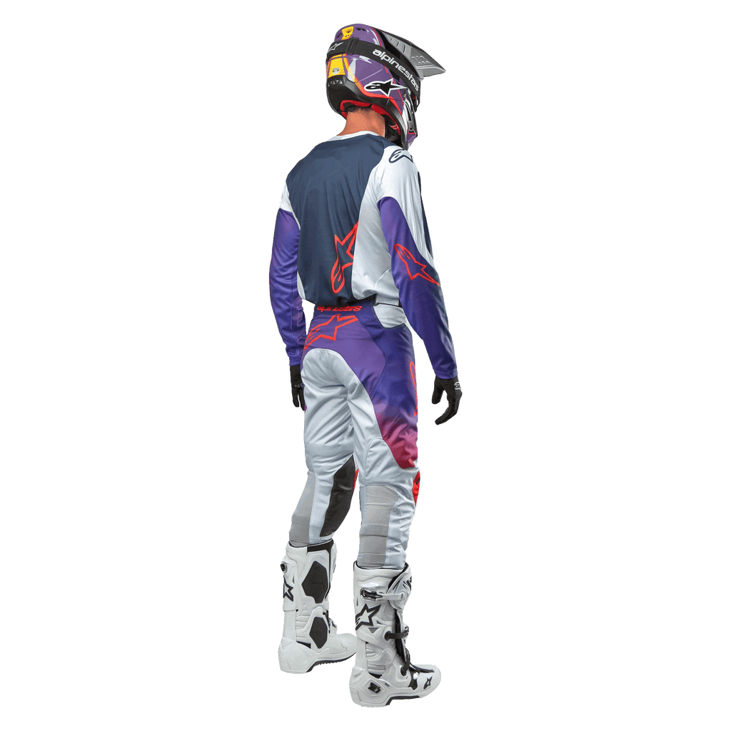 Alpinestars 2024 Racer Hoen Hose, Motocross-Ausrüstung, Rückansicht eines Fahrers in voller Montur, Hellgrau, Hot Orange und Schwarz mit violetten Akzenten, sportliches Design für Offroad-Rennen, inklusive Helm, Jersey und weißen Stiefeln