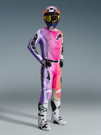 Pilote de motocross portant une tenue de course complète comprenant le pantalon Alpinestars 2026 Racer Air Portl, design multicolore rose, violet et orange vif avec accents blancs, maillot à manches longues assorti, casque noir avec lunettes jaunes et bottes blanches, tenue coordonnée pour la course tout-terrain haute performance.