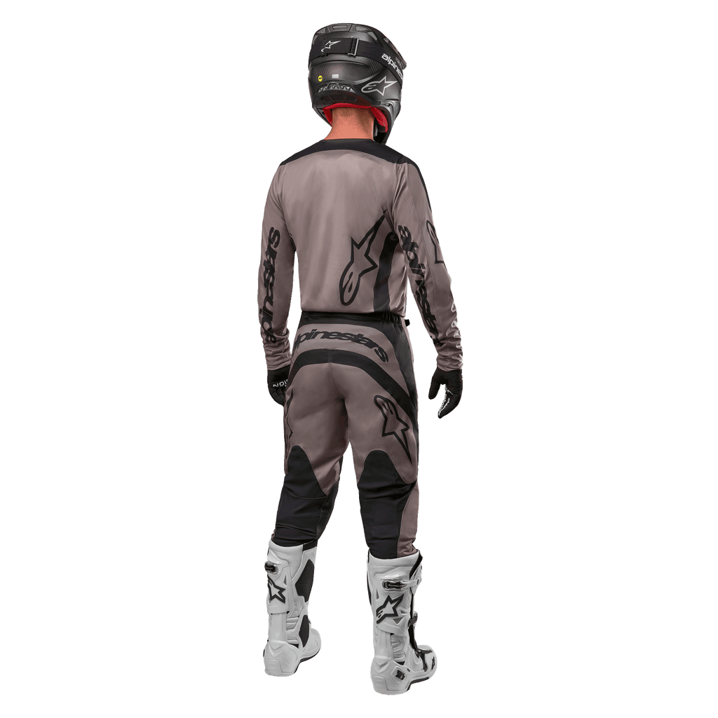 Alpinestars 2024 Fluid Lurv Trikot, Motocross-Trikot, Mud Black, Rückansicht eines Motocross-Fahrers in passender Rennausrüstung, schwarzer Helm und weiße Stiefel, markante schwarze Alpinestars-Logos, sportliche Passform für Offroad-Performance.