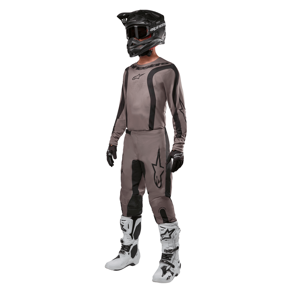 Motocross-Fahrer in kompletter Rennausrüstung mit dem Alpinestars 2024 Fluid Lurv Jersey in Mud Black, kombiniert mit der passenden Hose, schwarzem Helm mit Brille, weiß-schwarzen Stiefeln und schwarzen Handschuhen, koordiniertes Outfit für den Offroad-Rennsport