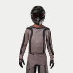 Motocross-Fahrer im Alpinestars 2024 Fluid Lurv Jersey in Mud Black, ein erdiger grau-brauner Stoff mit schwarzen Grafikstreifen und einem Stern-Logo auf der Brust, kombiniert mit schwarzem Helm, roter Brille und schwarzen Handschuhen für einen koordinierten Offroad-Rennlook