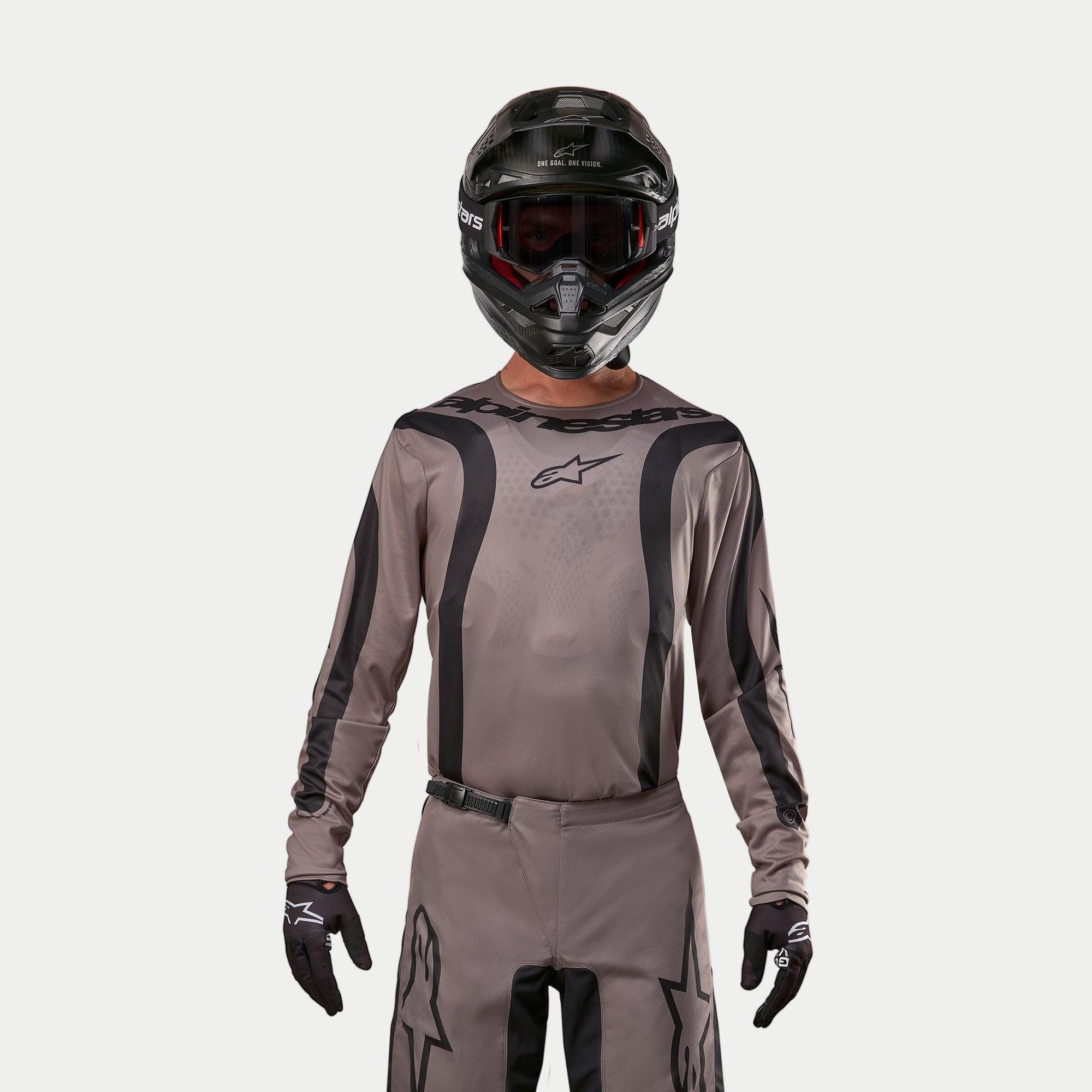 Motocross-Fahrer im Alpinestars 2024 Fluid Lurv Jersey in Mud Black, ein erdiger grau-brauner Stoff mit schwarzen Grafikstreifen und einem Stern-Logo auf der Brust, kombiniert mit schwarzem Helm, roter Brille und schwarzen Handschuhen für einen koordinierten Offroad-Rennlook