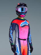 Alpinestars Limited Edition Supertech Records Jersey, motocross shirt en bijpassende uitrusting, meerkleurig met marineblauwe, roze en oranje spetteraccenten, groot blauw en wit logo op de borst, gedragen met bijpassende helm en handschoenen, atletische racepasvorm