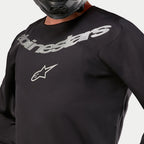 Alpinestars 2024 Fluid Graphite Jersey, Motocross Jersey, Black and Silver, close-up detail of the chest area featuring silver Alpinestars branding and star logo, breathable poly-fabric construction, worn by a rider in a black helmet