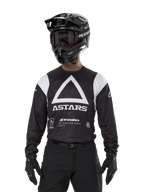 Maillot Alpinestars Techdura, noir, arborant un grand motif triangulaire blanc sur la poitrine avec le logo ASTARS et des empiècements blancs aux épaules, porté par un pilote avec un casque de motocross noir, des lunettes et une tenue tout-terrain noire assortie.