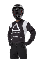 Maillot Alpinestars Techdura, maillot de motocross, noir et blanc, porté avec casque et gants assortis, pose les mains sur les hanches sur fond blanc, avec un grand logo A triangulaire et le marquage ASTARS