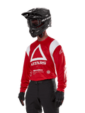 Alpinestars Techdura Jersey, motorcross jersey, helderrood en wit, met een opvallend wit driehoekontwerp en ASTARS-branding, gedragen met een zwarte helm en handschoenen, duurzame en vochtafvoerende off-road racekleding