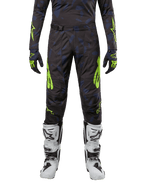 Alpinestars Techstar Rantera Pants 2024, pantalon de motocross, noir, bleu marine et jaune fluo, porté avec un maillot à manches longues, des gants assortis et des bottes de motocross blanches, avec des accents jaune fluo vifs et un motif sombre abstrait.