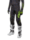 Pantalon Alpinestars Techstar Rantera 2024, pantalon de motocross, noir, bleu marine et jaune fluo, avec motifs camouflage bleu marine et logos vert néon, porté avec des gants assortis et des bottes blanches, coupe racing durable pour la performance tout-terrain