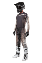 Pilote de motocross portant un équipement de course complet, maillot Techstar Pneuma 2024 en sable foncé et gris, pantalon de motocross à motifs assorti, casque noir avec lunettes, bottes blanches et noires, gants noirs, tenue coordonnée pour la course tout-terrain