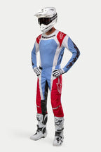 Pilote de motocross portant un équipement de course complet comprenant le maillot Alpinestars 2024 Techstar Ocuri en bleu clair, rouge Mars et blanc, associé à un pantalon assorti, un casque blanc avec des lunettes, des bottes blanches et des gants blancs et noirs.