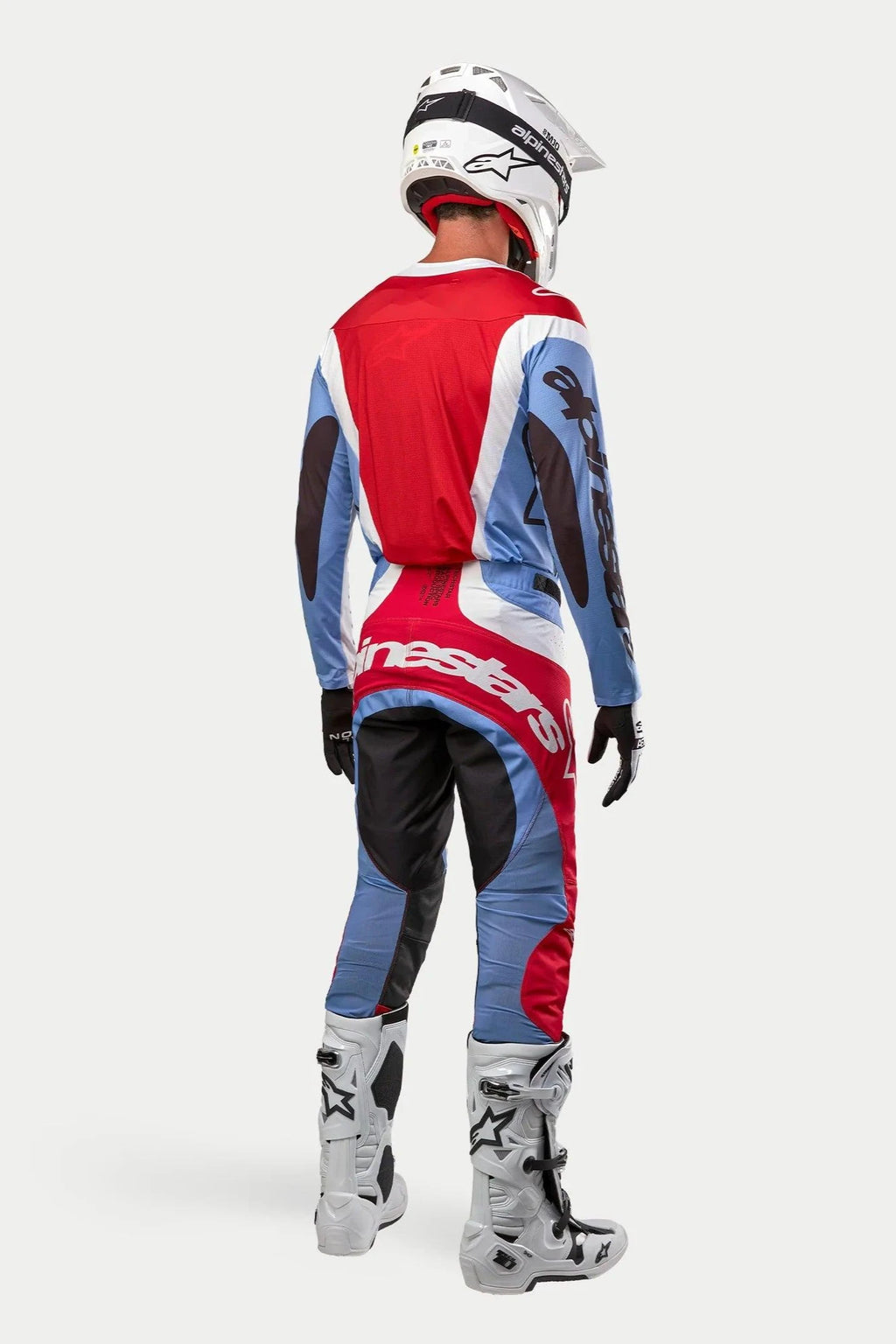 Pilote de motocross vu de dos portant un équipement de course complet, maillot Techstar Ocuri 2024 en bleu clair, rouge Mars et blanc, pantalon assorti avec marquage imposant, bottes blanches et noires, casque blanc, tenue coordonnée pour la course tout-terrain