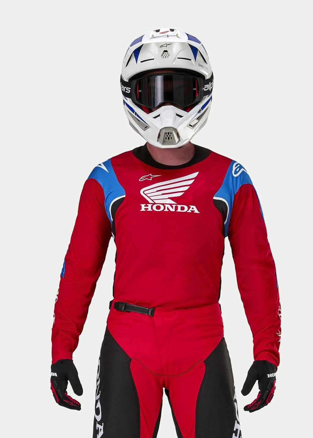 Motocross-Fahrer im Alpinestars Honda Racer Iconic Trikot in leuchtendem Rot mit blauen und weißen Akzenten sowie Honda-Branding, kombiniert mit einem weiß-blauen Helm, dunkler Brille und passender rot-schwarzer Motocross-Hose, Performance-Ausrüstung für den Offroad-Einsatz