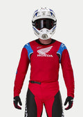Motocross-Fahrer im Alpinestars Honda Racer Iconic Trikot in leuchtendem Rot mit blauen und weißen Akzenten sowie Honda-Branding, kombiniert mit einem weiß-blauen Helm, dunkler Brille und passender rot-schwarzer Motocross-Hose, Performance-Ausrüstung für den Offroad-Einsatz