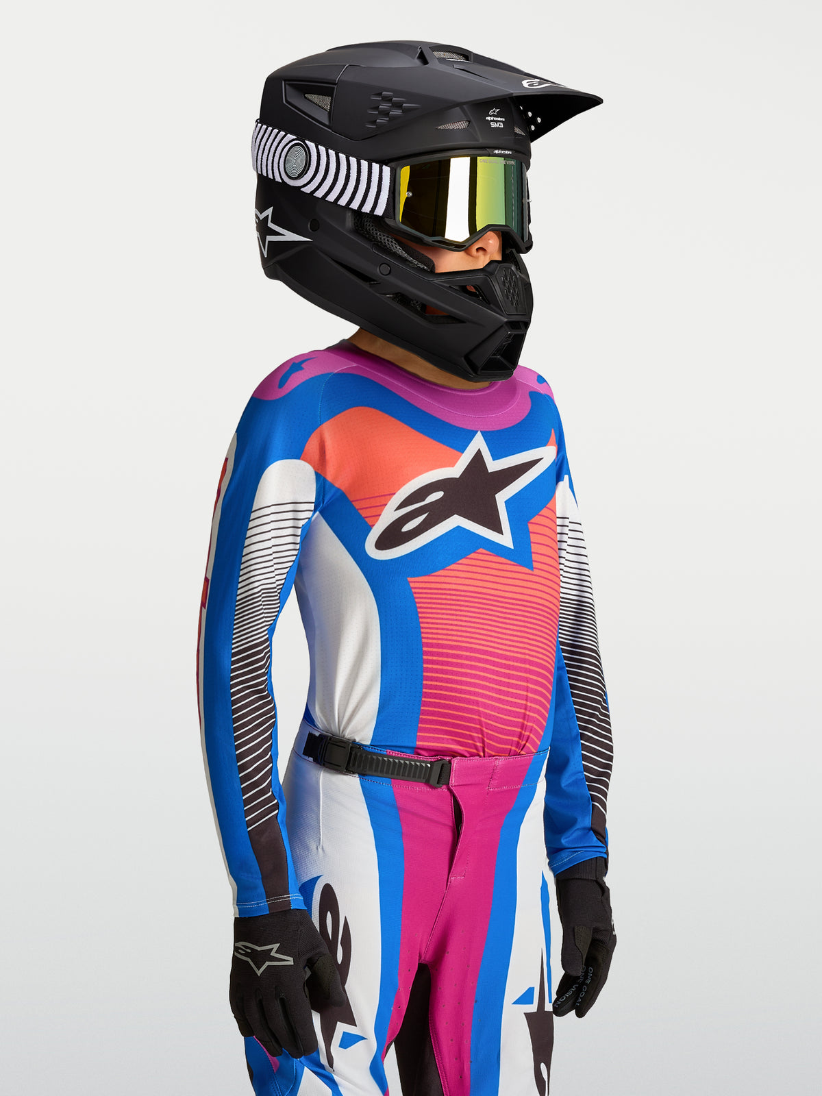 Alpinestars Supertech Jugend-Trikot Maven 2027, atmungsaktives Motocross-Trikot für junge Fahrer, Blau und Koralle, kombiniert mit schwarzem Rennhelm und Schutzausrüstung.