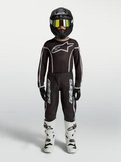 Alpinestars jeugd Fluid Apex V2 jersey en gear set, zwart-zilveren motocross-outfit met witte motorlaarzen en helm.