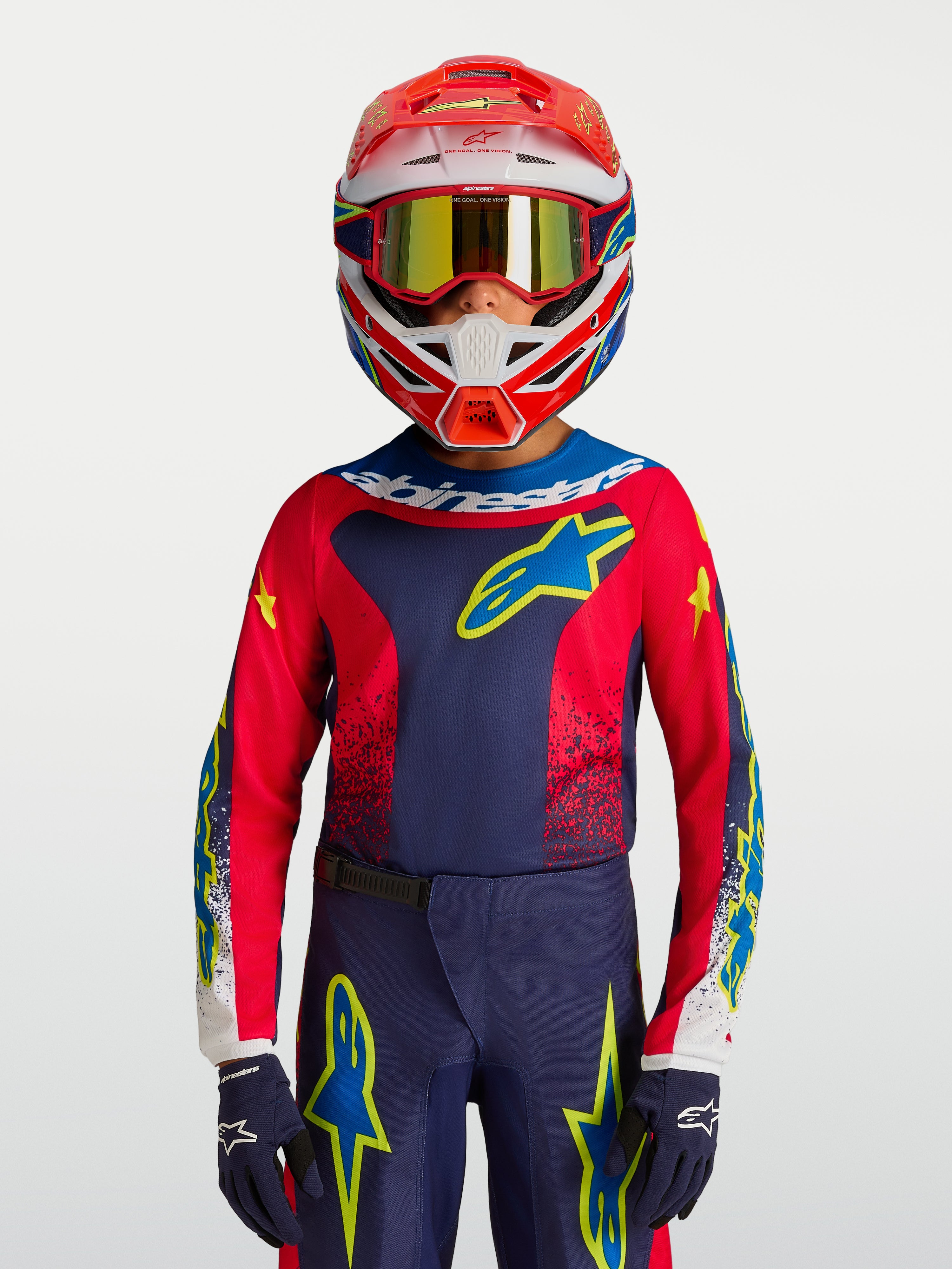 Maillot Fluid Drag Junior 2027 d'Alpinestars, maillot de motocross pour enfant, bleu marine et rouge avec accents jaunes, porté par un jeune pilote avec casque et gants.