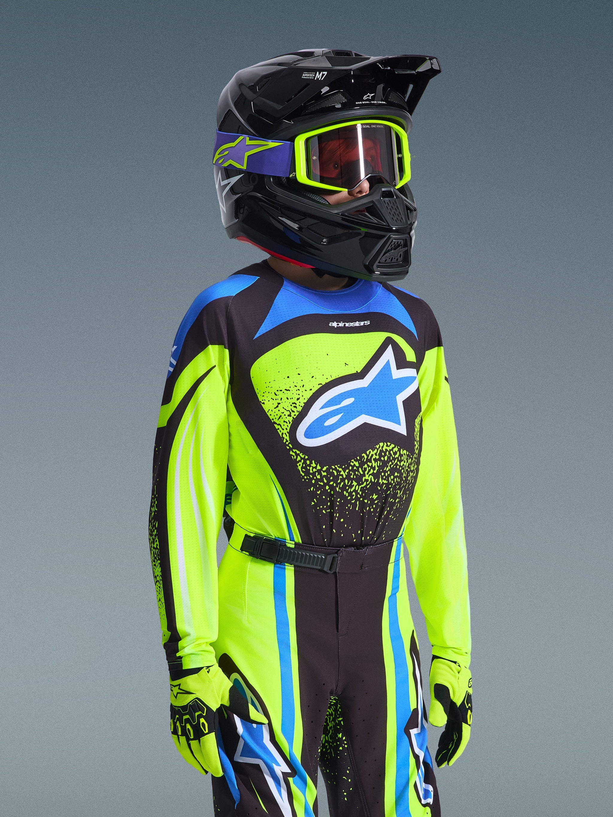 Jeugd motorcrossrijder in Alpinestars 2026 Youth Supertech Nomur Jersey in Yellow Fluo en Dark Navy, met een blauw Astars-logo, bijpassende racebroek en handschoenen, en een zwarte helm met fluo gele bril, professionele off-road motorcrossuitrusting.