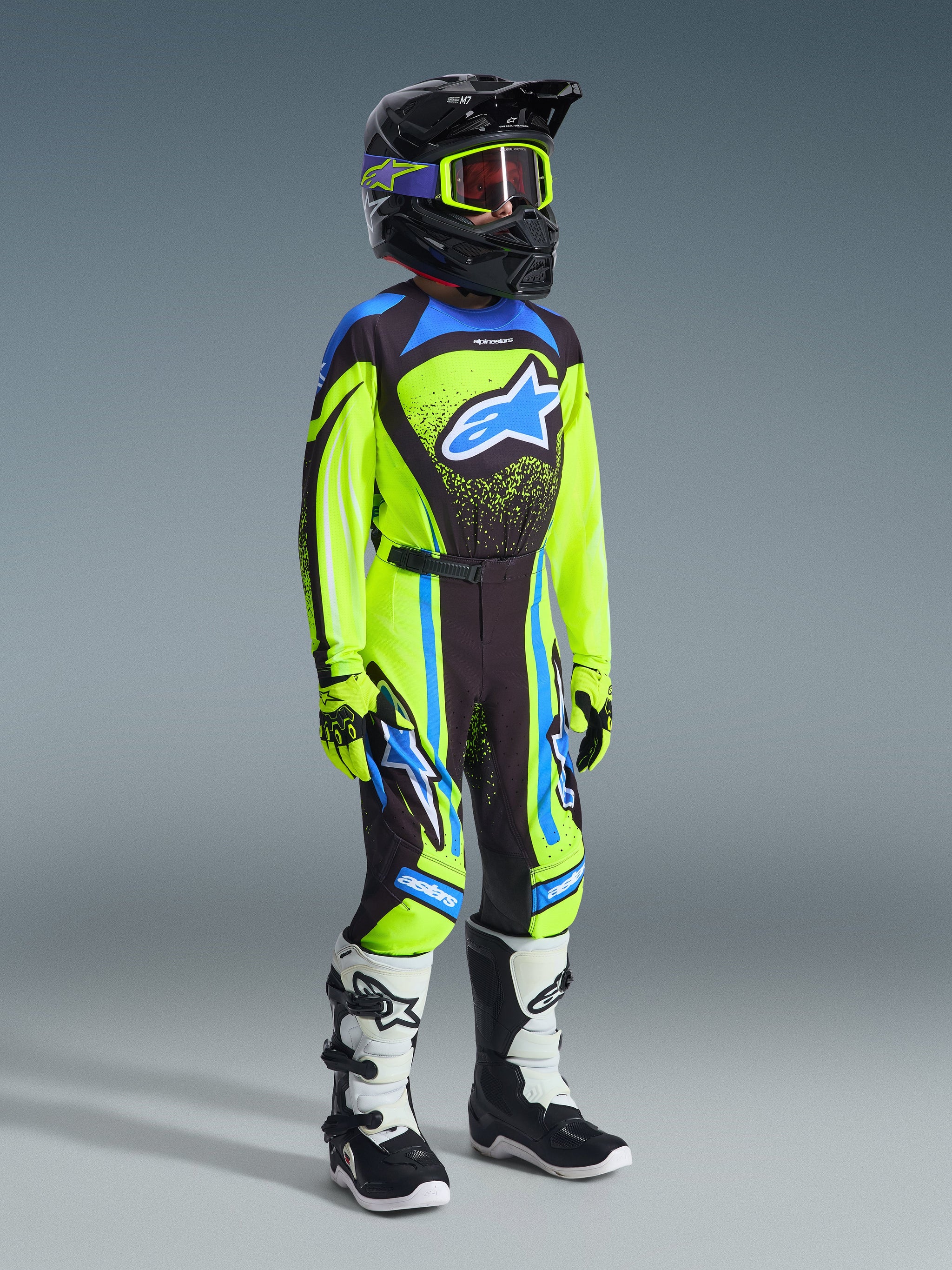 Jeugd motocrossrijder in complete race-uitrusting, 2026 Youth Supertech Nomur Jersey in Yellow Fluo en Dark Navy, bijpassende broek met blauwe accenten, zwarte helm met fluogele crossbril, witte en zwarte laarzen, gecoördineerde outfit voor off-road racen.