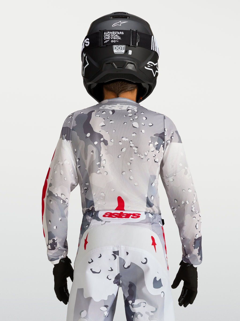 Alpinestars Maillot Fluid Lone Junior 2027, maillot de motocross pour enfant, vue de dos, gris et blanc avec motif camouflage et accents rouges, porté avec un casque noir, équipement off-road haute performance