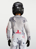 Alpinestars Maillot Fluid Lone Junior 2027, maillot de motocross pour enfant, vue de dos, gris et blanc avec motif camouflage et accents rouges, porté avec un casque noir, équipement off-road haute performance
