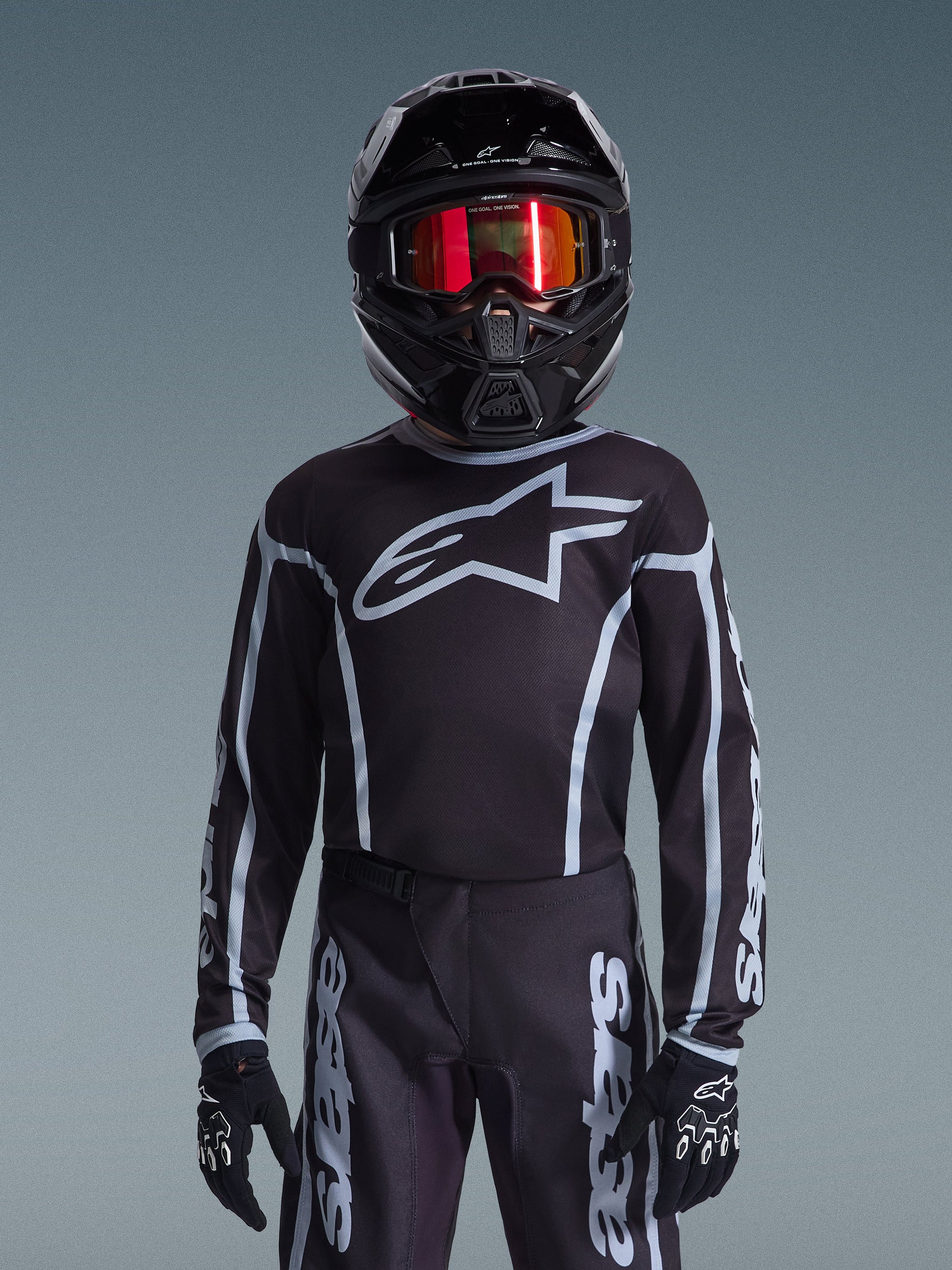 Jonge motorcrosser in Alpinestars 2026 Jeugd Fluid Apex Jersey in zwart en grijs, bijpassende broek, zwarte helm met rode reflecterende crossbril en zwarte handschoenen, gecoördineerde off-road race-uitrusting.