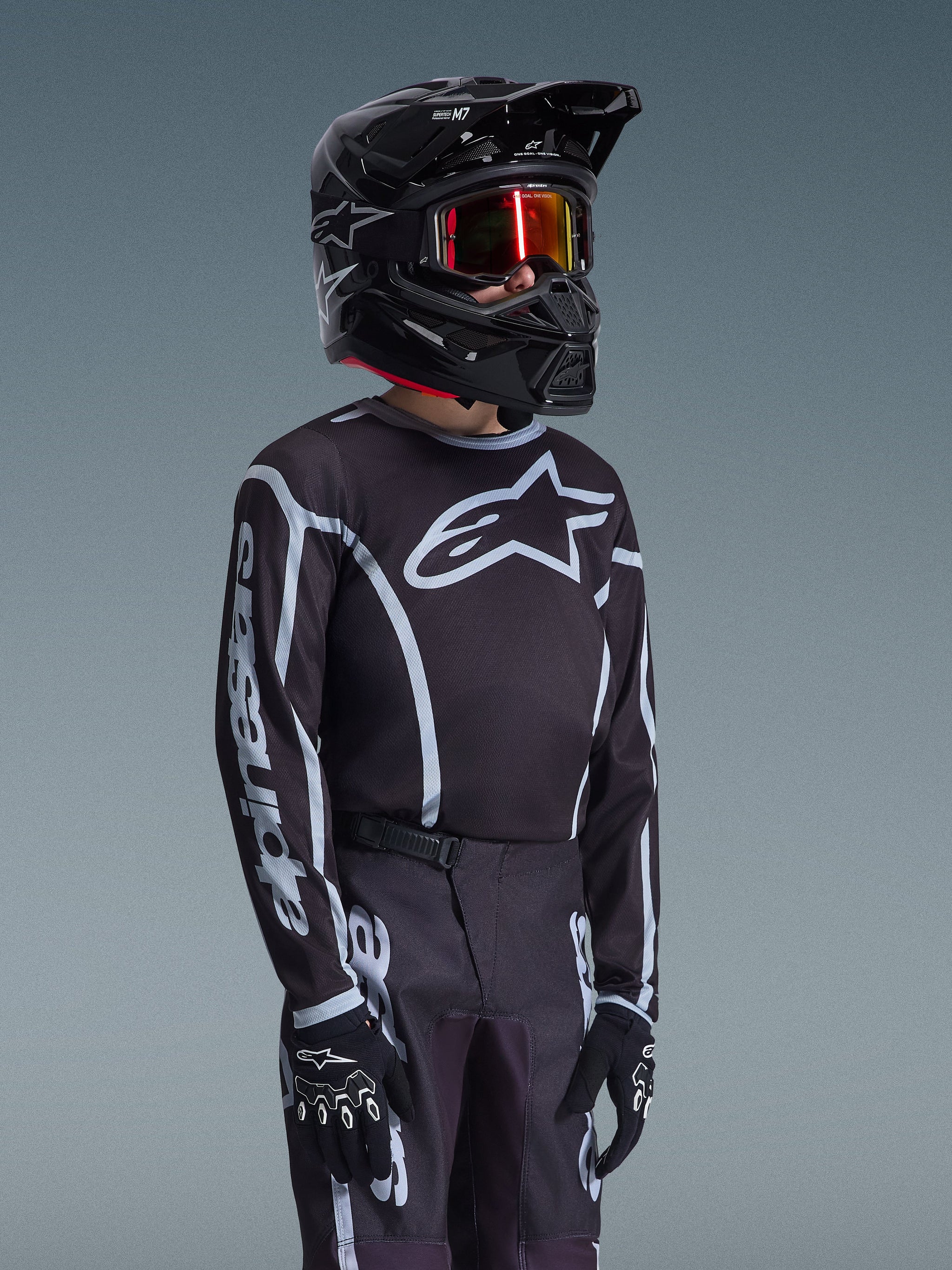 Alpinestars 2026 Youth Fluid Apex Jersey in zwart en grijs, gedragen door een jonge rijder met een bijpassende zwarte helm en getinte bril, met opvallende Alpinestars-branding op de borst en mouwen, onderdeel van een gecoördineerde motorcross-uitrusting.