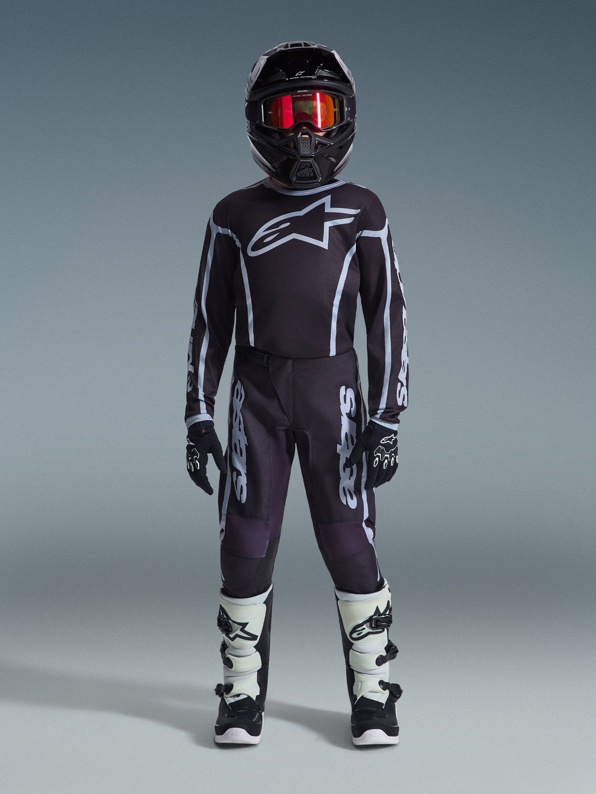 Jonge motorcrossrijder in de Alpinestars 2026 Youth Fluid Apex Jersey in zwart en grijs, gecombineerd met een bijpassende broek, zwarte helm met rood iriserende bril, zwart-witte handschoenen en motorcrosslaarzen, complete uitrustingsset voor jeugd off-road racen