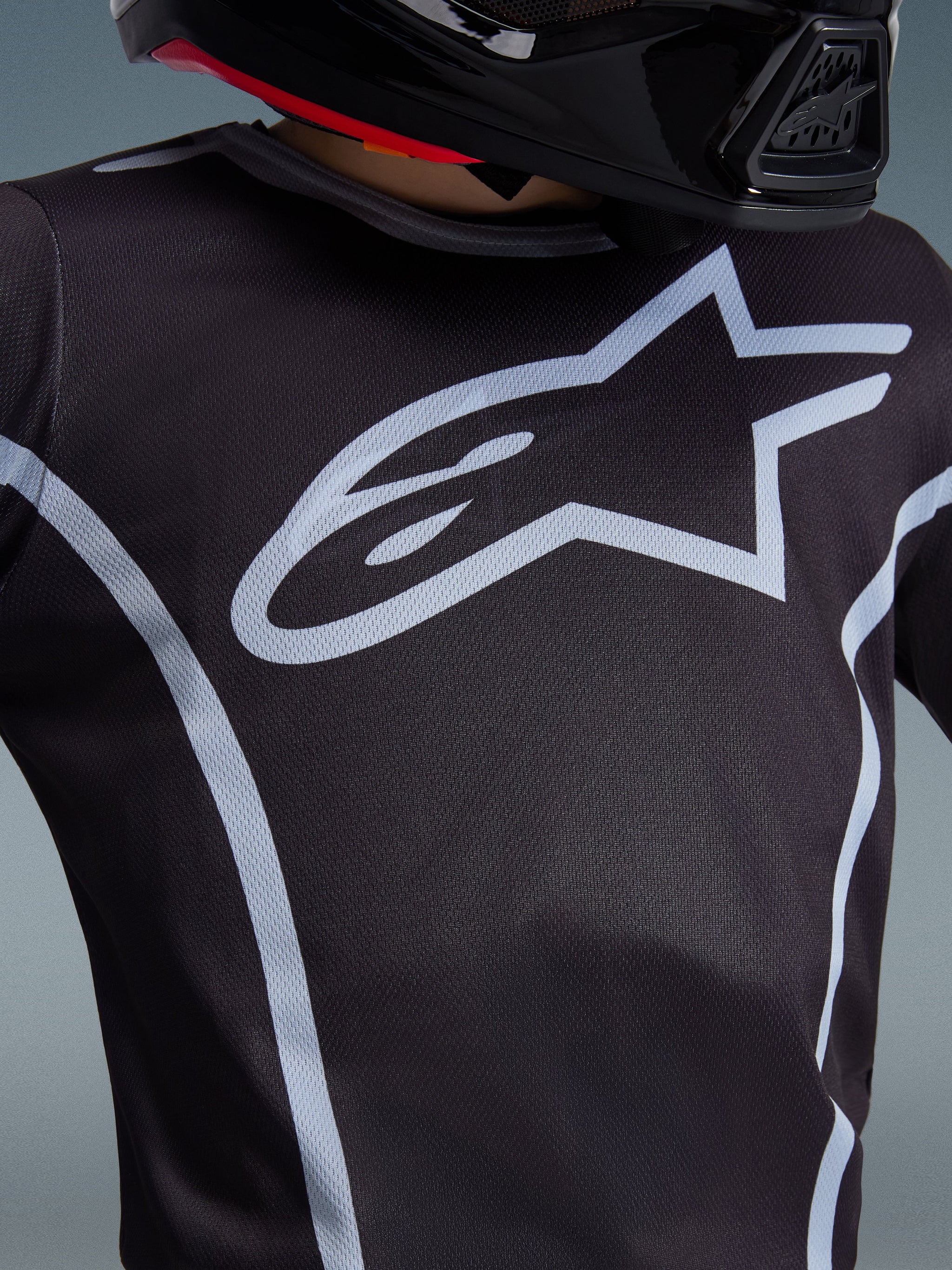 Alpinestars 2026 Youth Fluid Apex Jersey, detailopname van het borstontwerp, zwart en grijs, met een opvallend lichtgrijs sterlogo en sportieve biezen, gedragen met een zwarte helm, performance motorcrossuitrusting voor jonge rijders