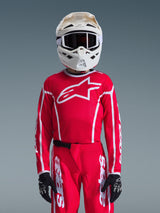 Maillot Alpinestars Youth Fluid Apex 2026, rouge et blanc, porté avec un pantalon de motocross assorti, un casque blanc avec des lunettes noires et des gants noirs, arborant un grand logo blanc et une coupe athlétique pour les courses tout-terrain junior.