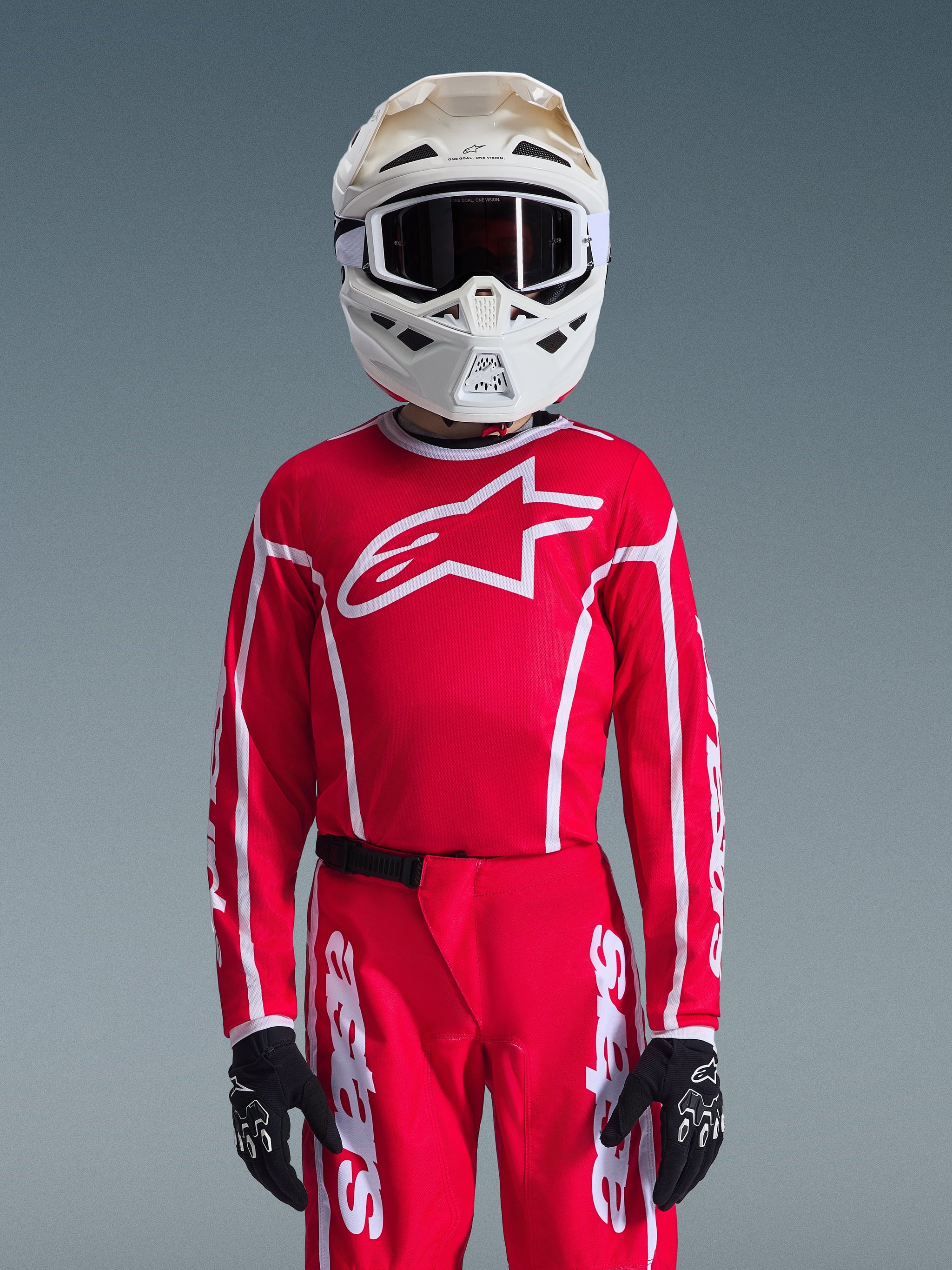 Maillot Alpinestars Youth Fluid Apex 2026, rouge et blanc, porté avec un pantalon de motocross assorti, un casque blanc avec des lunettes noires et des gants noirs, arborant un grand logo blanc et une coupe athlétique pour les courses tout-terrain junior.
