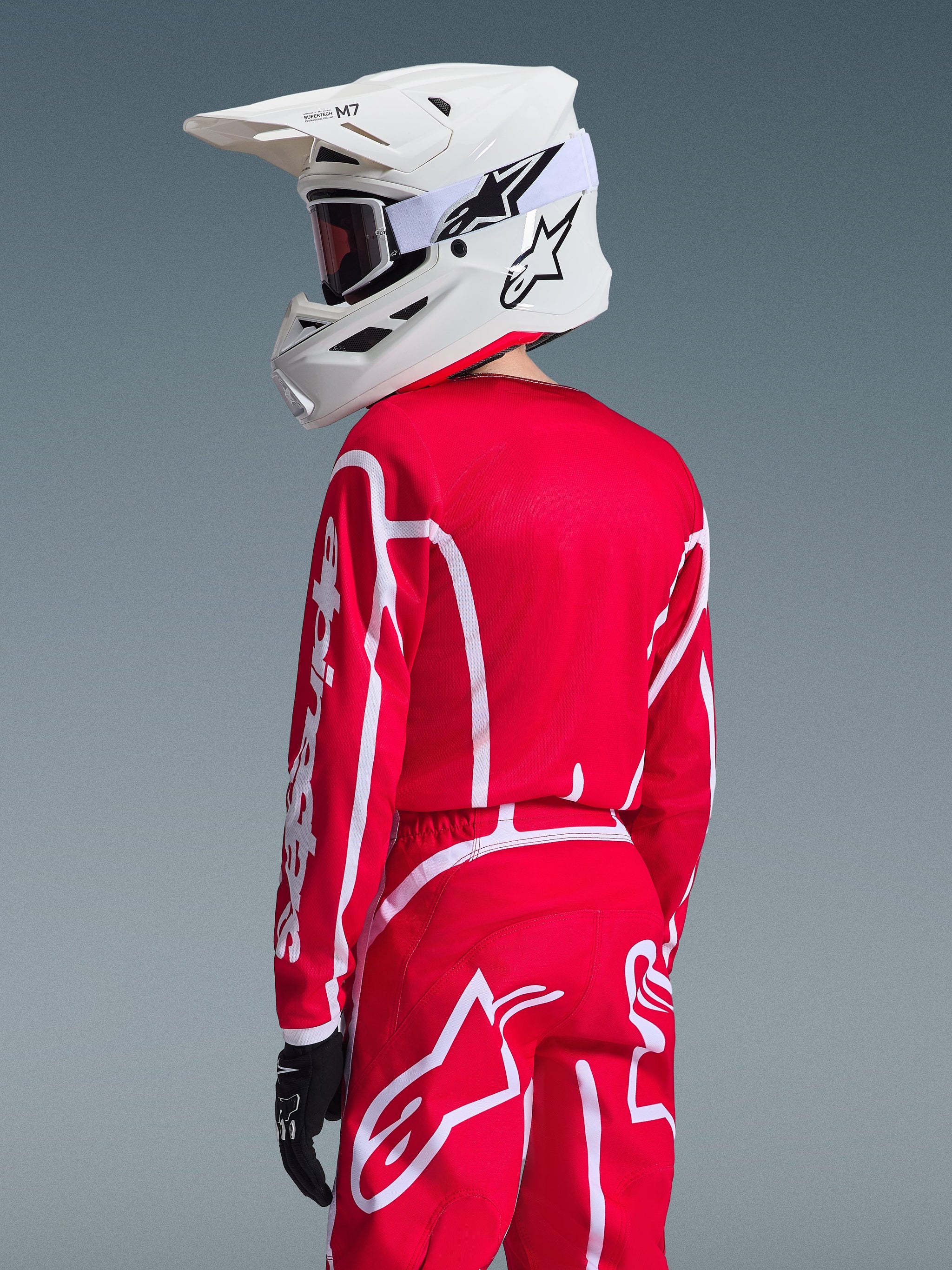 Maillot Alpinestars Youth Fluid Apex 2026 rouge et blanc, maillot de motocross pour enfant porté avec une tenue assortie et un casque blanc, vue de dos montrant la coupe athlétique et le logo Alpinestars blanc sur la manche et le pantalon
