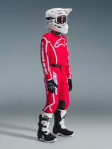 Jeune pilote de motocross portant une tenue de course complète, Maillot Junior Fluid Apex 2026 rouge avec logos Alpinestars blancs, pantalon rouge assorti avec graphismes blancs et accents noirs, casque blanc avec lunettes, bottes blanches et noires, gants noirs et blancs, tenue coordonnée pour les jeunes pilotes tout-terrain
