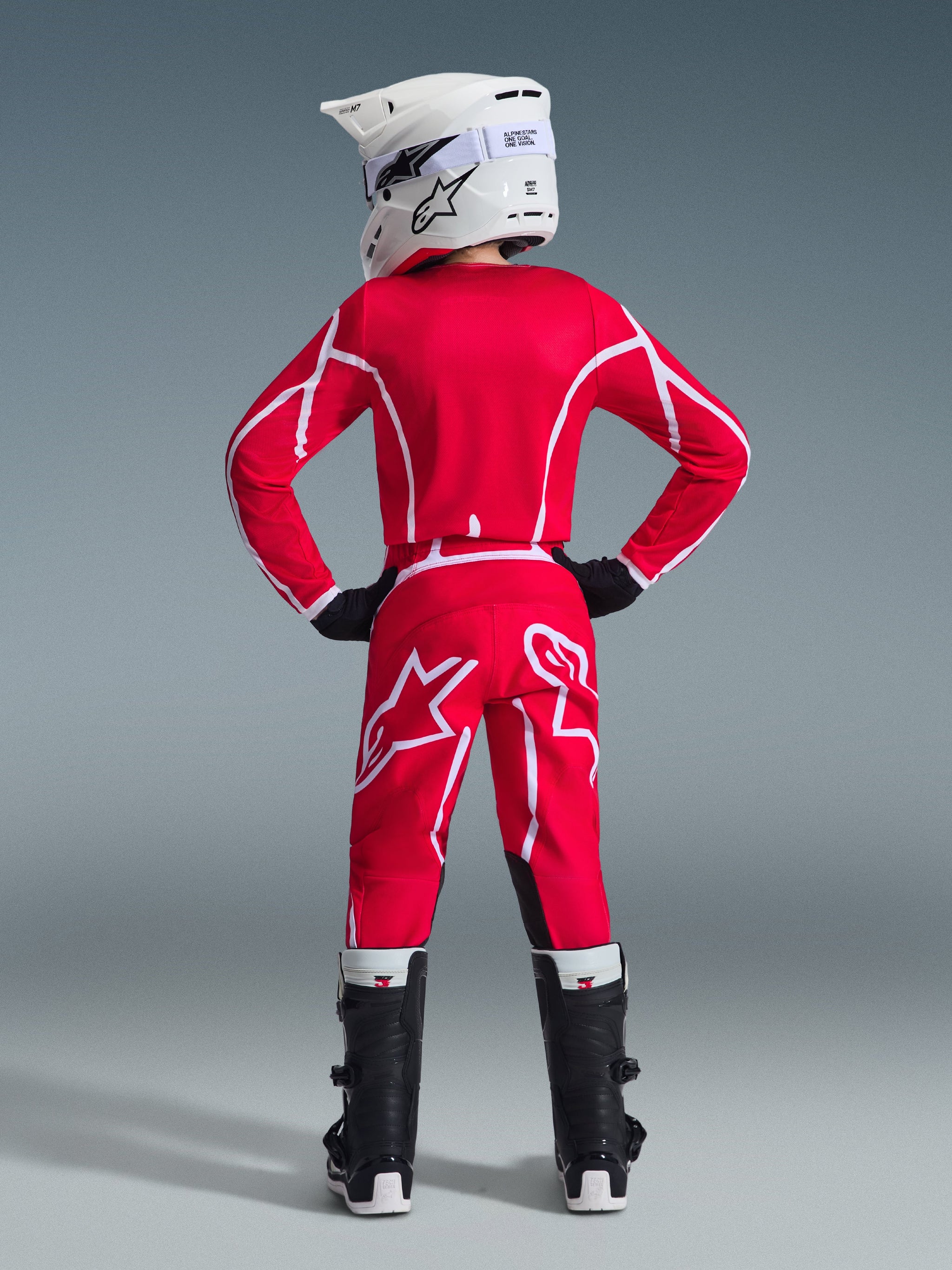 Vue arrière d'un jeune pilote de motocross portant le maillot Alpinestars Youth Fluid Apex 2026 rouge et blanc, pantalon assorti, casque blanc avec lunettes et bottes noires, tenue complète de course tout-terrain.