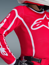 Gros plan sur le maillot Alpinestars Junior Fluid Apex 2026 rouge et blanc, arborant un large logo blanc sur le torse et des motifs géométriques sur les manches, porté par un pilote avec des gants de motocross noirs et un casque blanc.