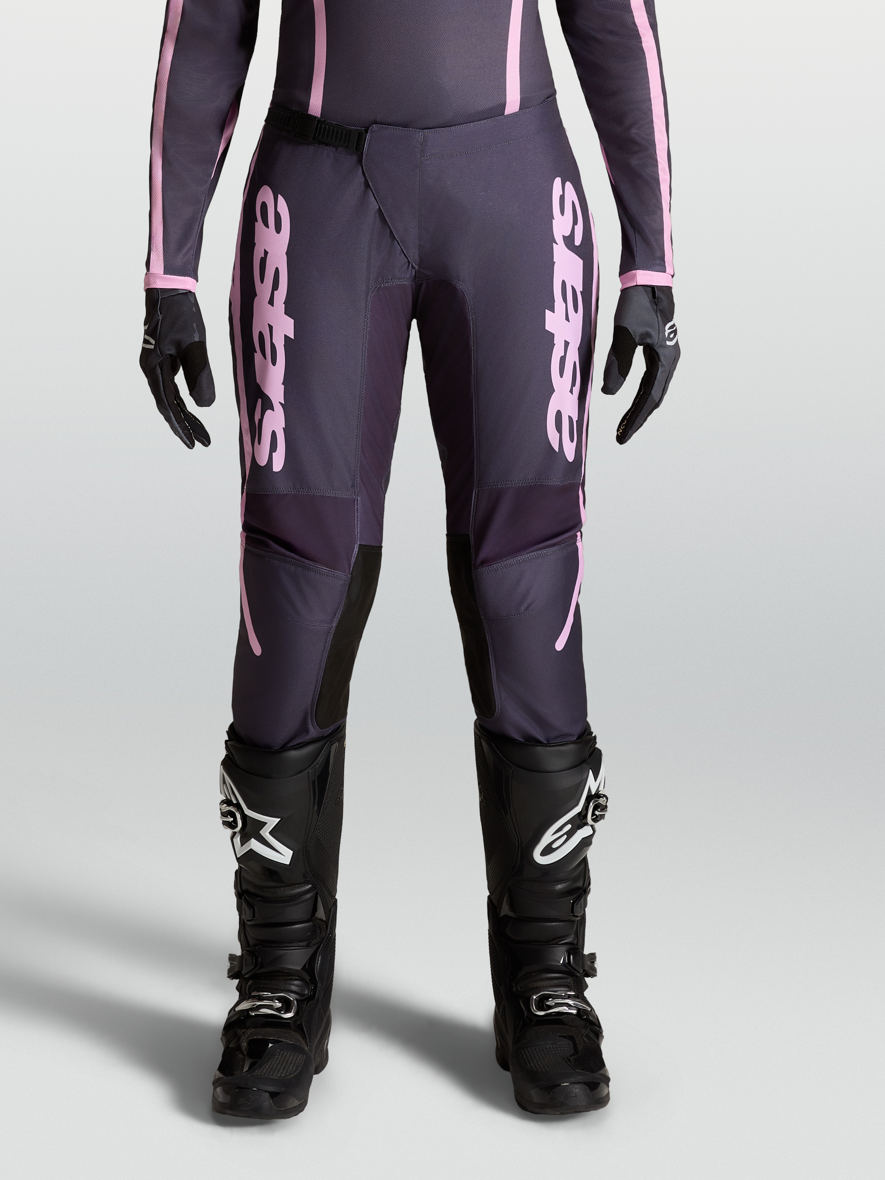 Alpinestars Stella Fluid Apex V2 Hose, Damen-Motocross-Hose in Grau und Pink, Modell an der Frontansicht mit Ratschenverschluss, 600D-Polygewebe, kombiniert mit schwarzen MX-Stiefeln und Handschuhen.