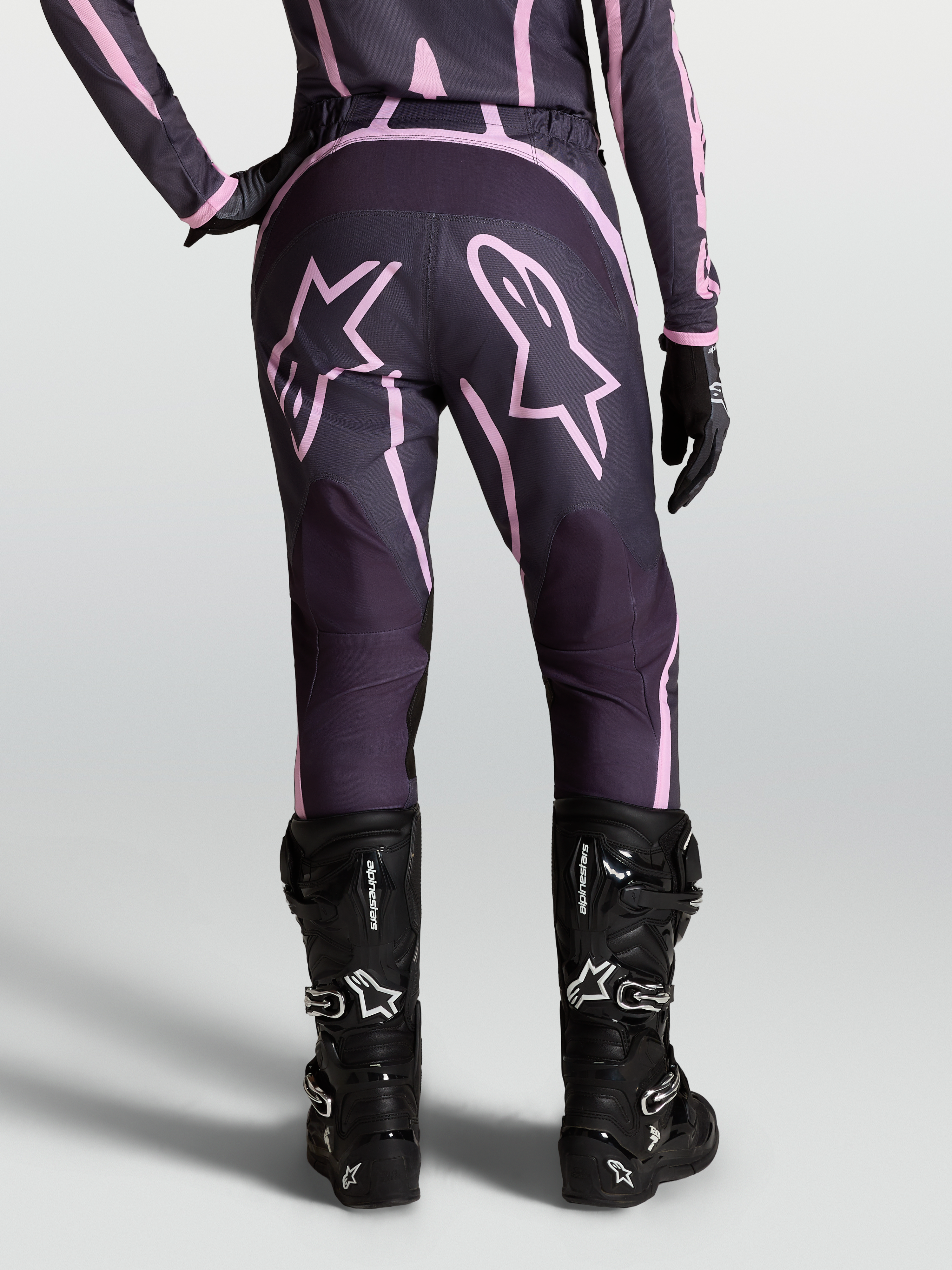 Alpinestars Stella Fluid Apex V2 broek, dames motorcrossbroek in grijs en roze met ster-graphics, gedragen met zwarte MX-laarzen en handschoenen.