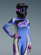 Pilote de motocross portant l'ensemble Alpinestars 2026 Stella Techstar Nomur, maillot bleu clair avec des accents roses et violets, pantalon bleu et blanc assorti, casque intégral noir avec lunettes violettes et gants roses et noirs, équipement de course tout-terrain professionnel pour femme.