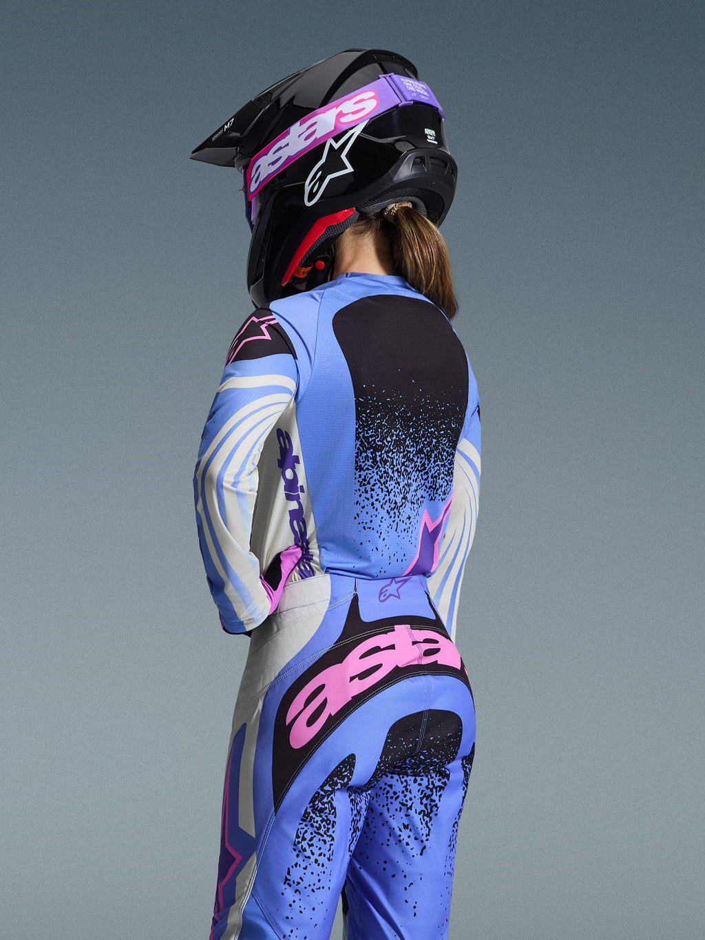 Vue arrière d'une pilote de motocross portant la tenue Alpinestars 2026 Stella Techstar Nomur, bleu clair avec des accents roses et noirs, maillot et pantalon assortis, casque noir avec lunettes roses, coupe ergonomique pour femme pour la course tout-terrain.