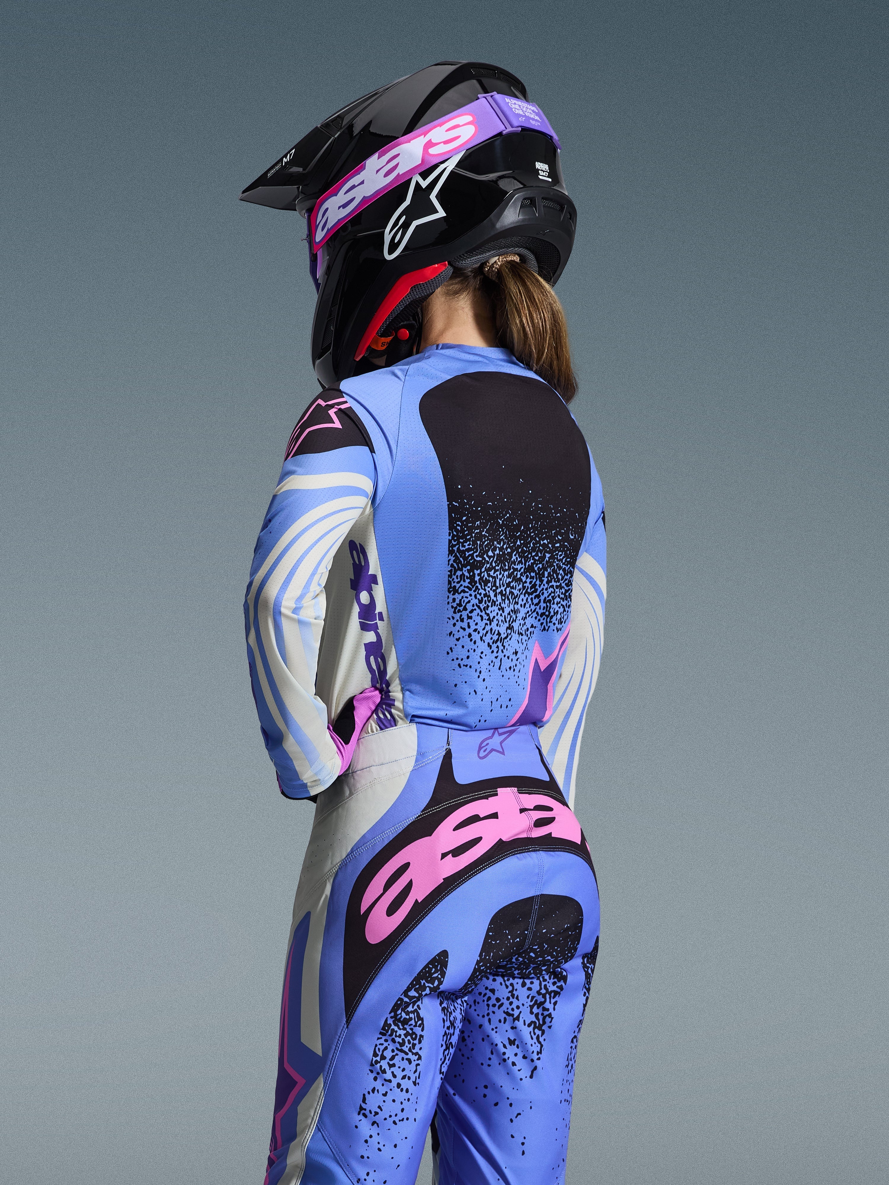 Vue arrière d'une pilote de motocross portant la tenue Alpinestars 2026 Stella Techstar Nomur, bleu clair avec des accents roses et noirs, maillot et pantalon assortis, casque noir avec lunettes roses, coupe ergonomique pour femme pour la course tout-terrain.