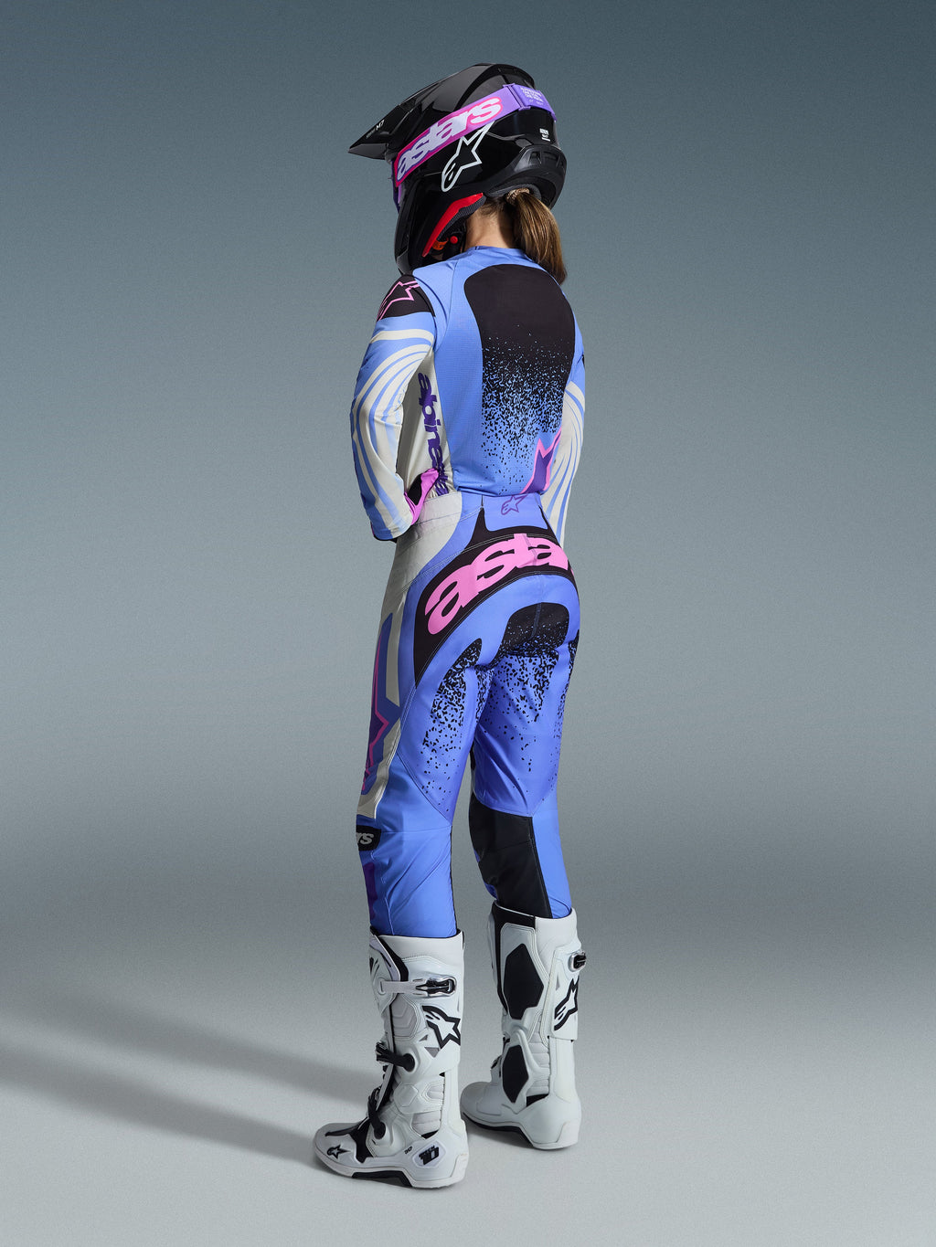 Alpinestars 2026 Stella Techstar Nomur Gear Set, vue arrière d'une pilote de motocross portant un ensemble maillot et pantalon bleu clair, blanc et rose avec des détails mouchetés noirs, assorti à un casque noir et des bottes blanches, équipement de course professionnel pour femmes.